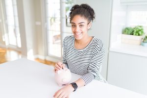 Young african american woman putting a coin inside piggy bank as invest or saving with a happy face standing and smiling with a confident smile showing teeth